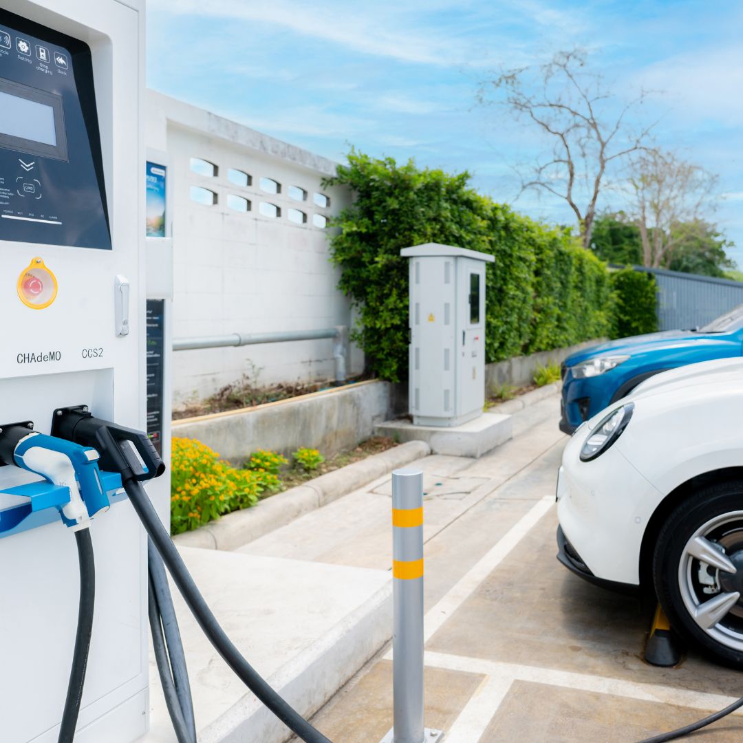 Alt text: "A contemporary convenience store with electric vehicle charging stations prominently displayed. The foreground showcases charging stations equipped with cables for connecting electric vehicles, while customers take advantage of this eco-friendly amenity. The store's entrance and colorful signage welcome visitors, hinting at the diverse range of products available inside. The charging stations reflect the store's commitment to sustainability and accessibility for electric vehicle owners."