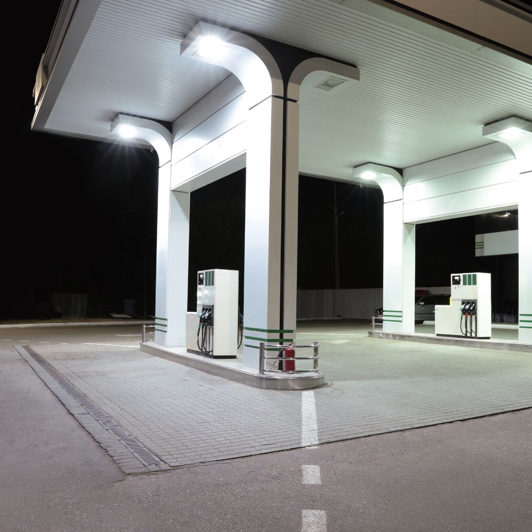 "An exterior view of a convenience store with well-lit surroundings. Outdoor lighting fixtures illuminate the parking lot and walkways, ensuring visibility and safety. The convenience store signage is prominently displayed and illuminated. Power meters and utility connections are visible on the side of the building, providing electrical supply to the store. Security cameras are mounted on the exterior walls, enhancing safety and surveillance. Customers can be seen entering and exiting the store, creating a bustling atmosphere."